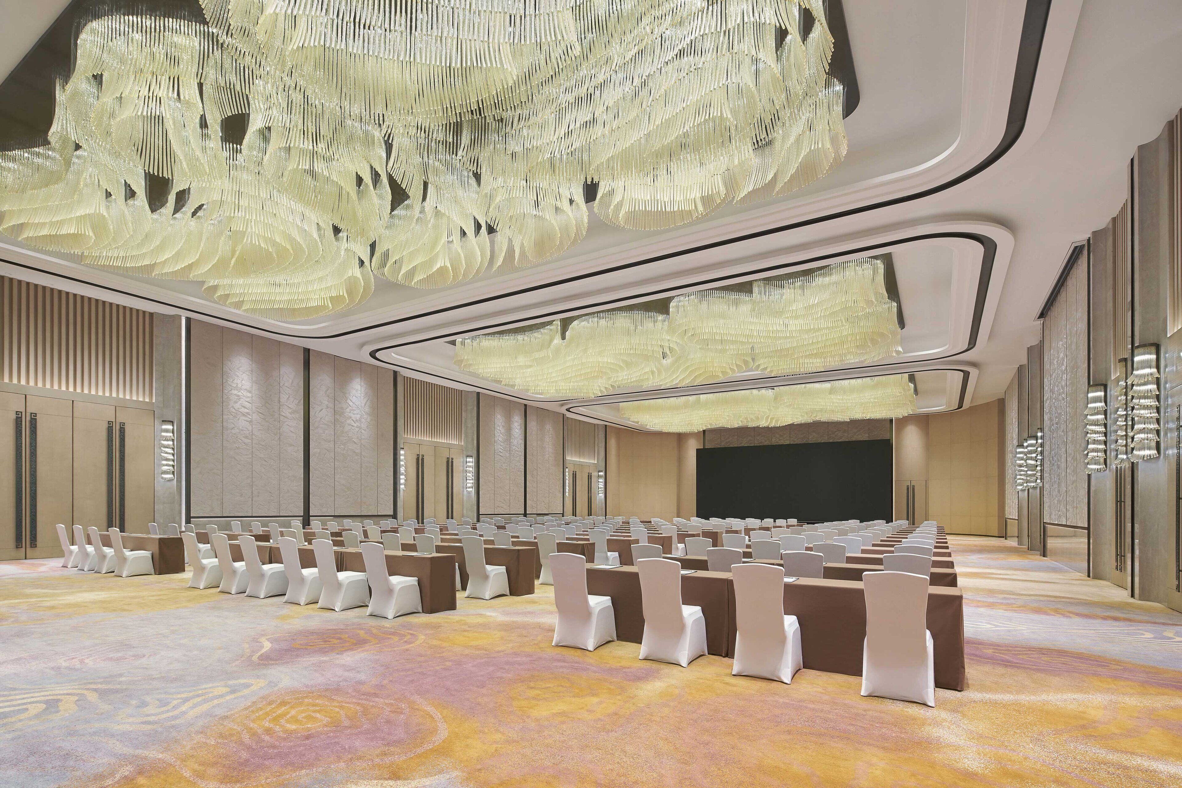 Une salle de conférence avec des rangées de chaises et de tables, un grand lustre et un écran à l’avant.