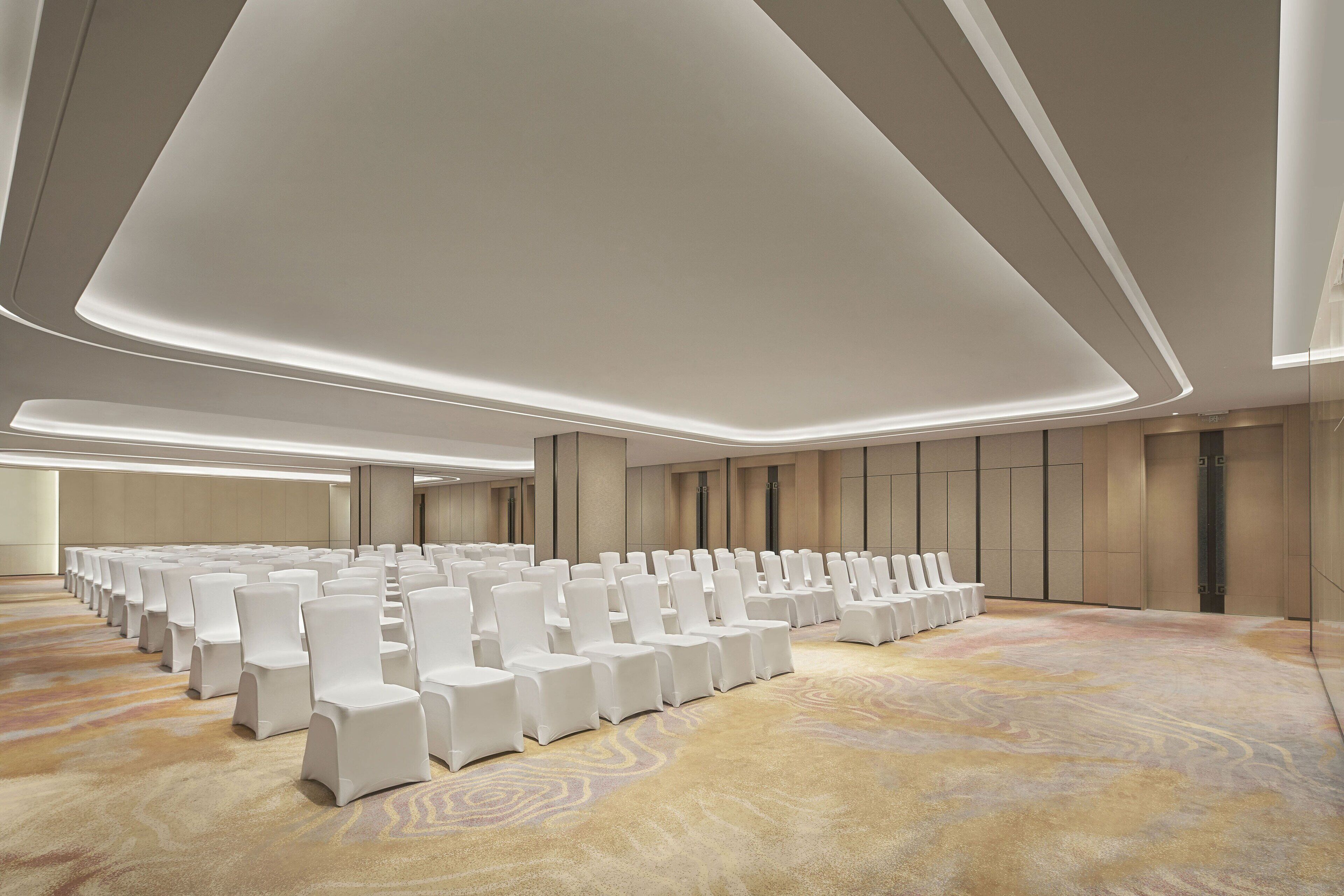 A conference room with rows of white chairs, a patterned carpet, and recessed ceiling lighting.