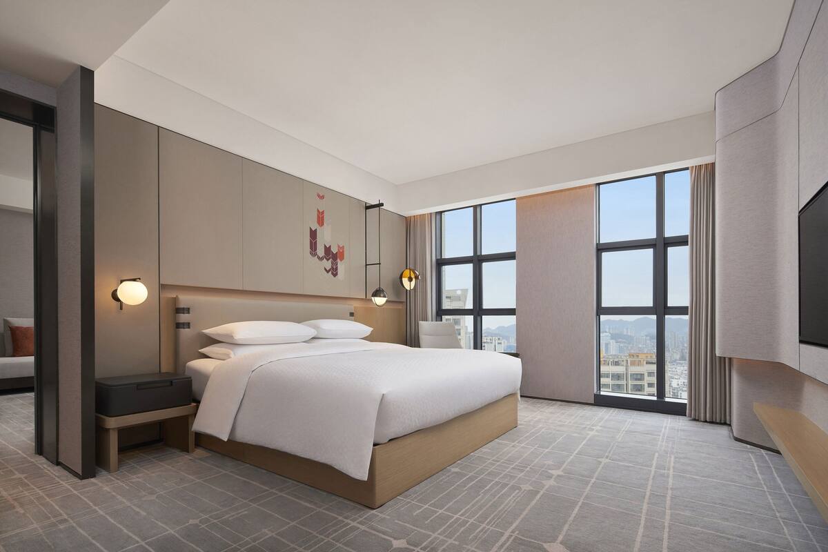 A modern hotel room with a large bed, a desk, a chair, a TV, and a view of the city through large windows.