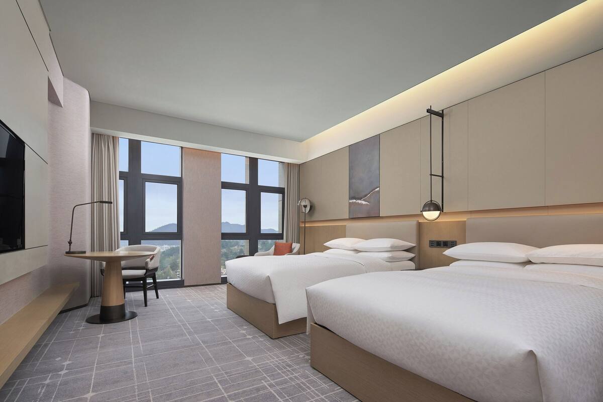 A hotel room with two beds, a desk, a chair, a large window, and a TV.