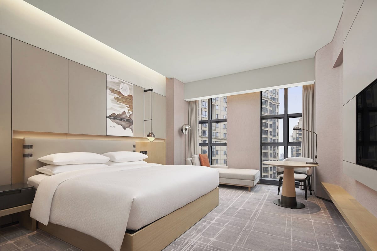 A modern hotel room with a large bed, a desk, and a view of the city through large windows.