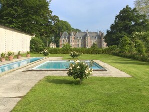 Une grande piscine rectangulaire, entourée d’une pelouse et bordée d’une haie. Au fond, on aperçoit un grand immeuble à plusieurs étages, de style architectural classique.