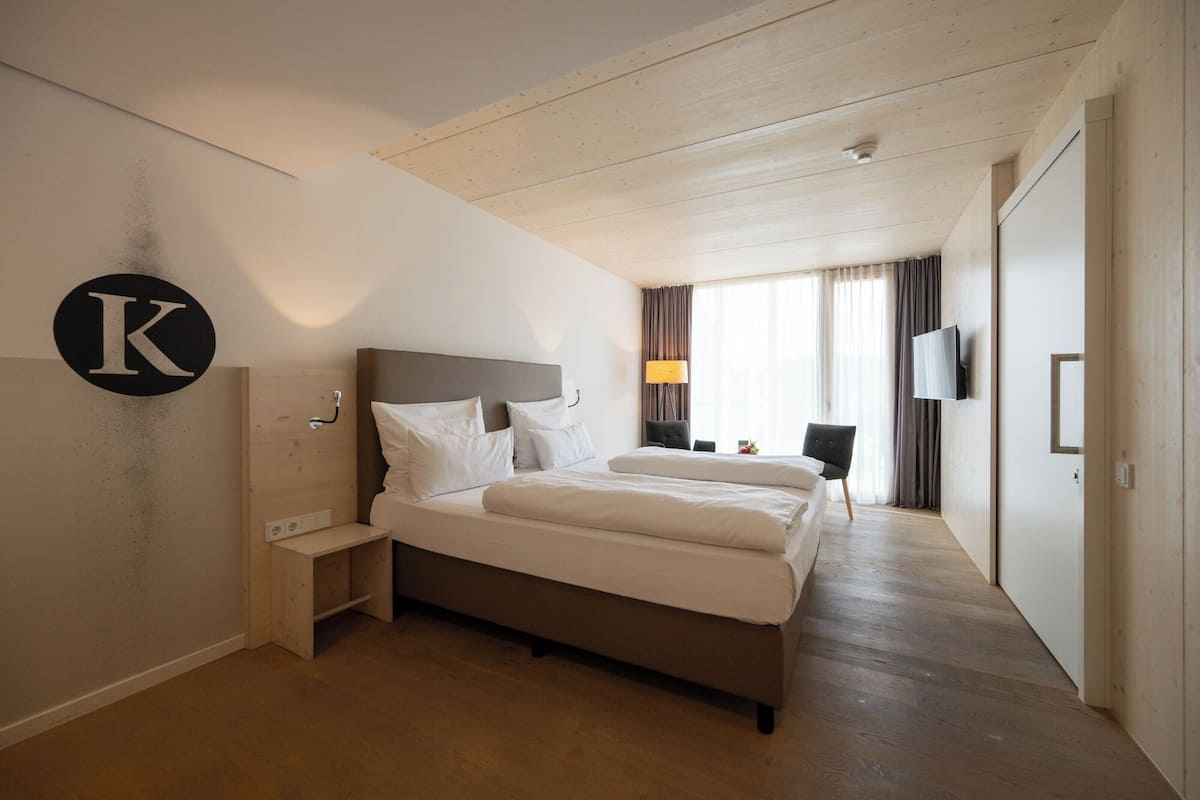 A hotel room with a large bed, a bedside table, a lamp, a TV, and a large window with curtains.