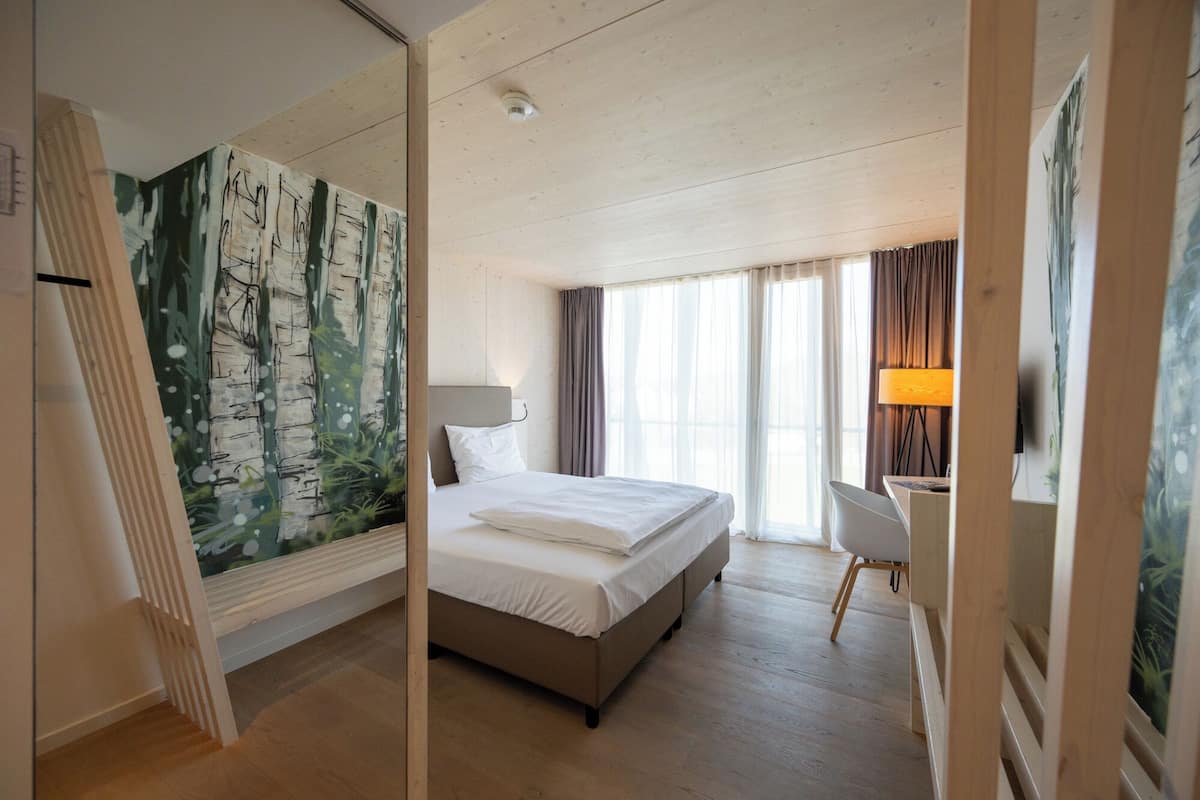 A hotel room with a bed, a desk, a chair, a lamp, and a large painting on the wall.
