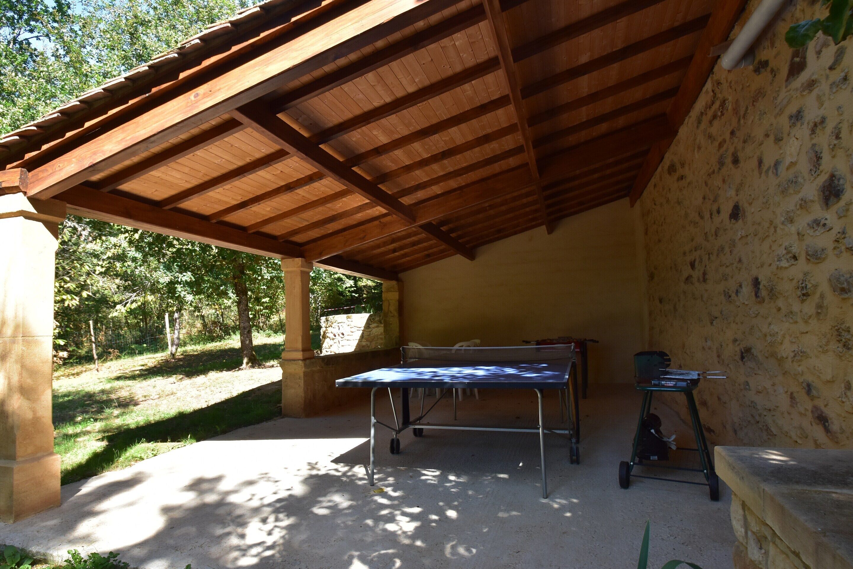An outdoor covered area with a table and grill, surrounded by stone walls and greenery.