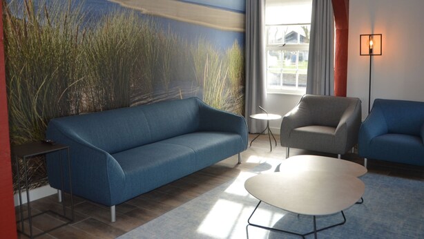 A modern living room with a blue sofa, a grey armchair, and a white coffee table. A painting of a beach scene is on the wall, and there is a window with curtains. - Sleeping in a National Monument is Possible on Texel (Den Hoorn)