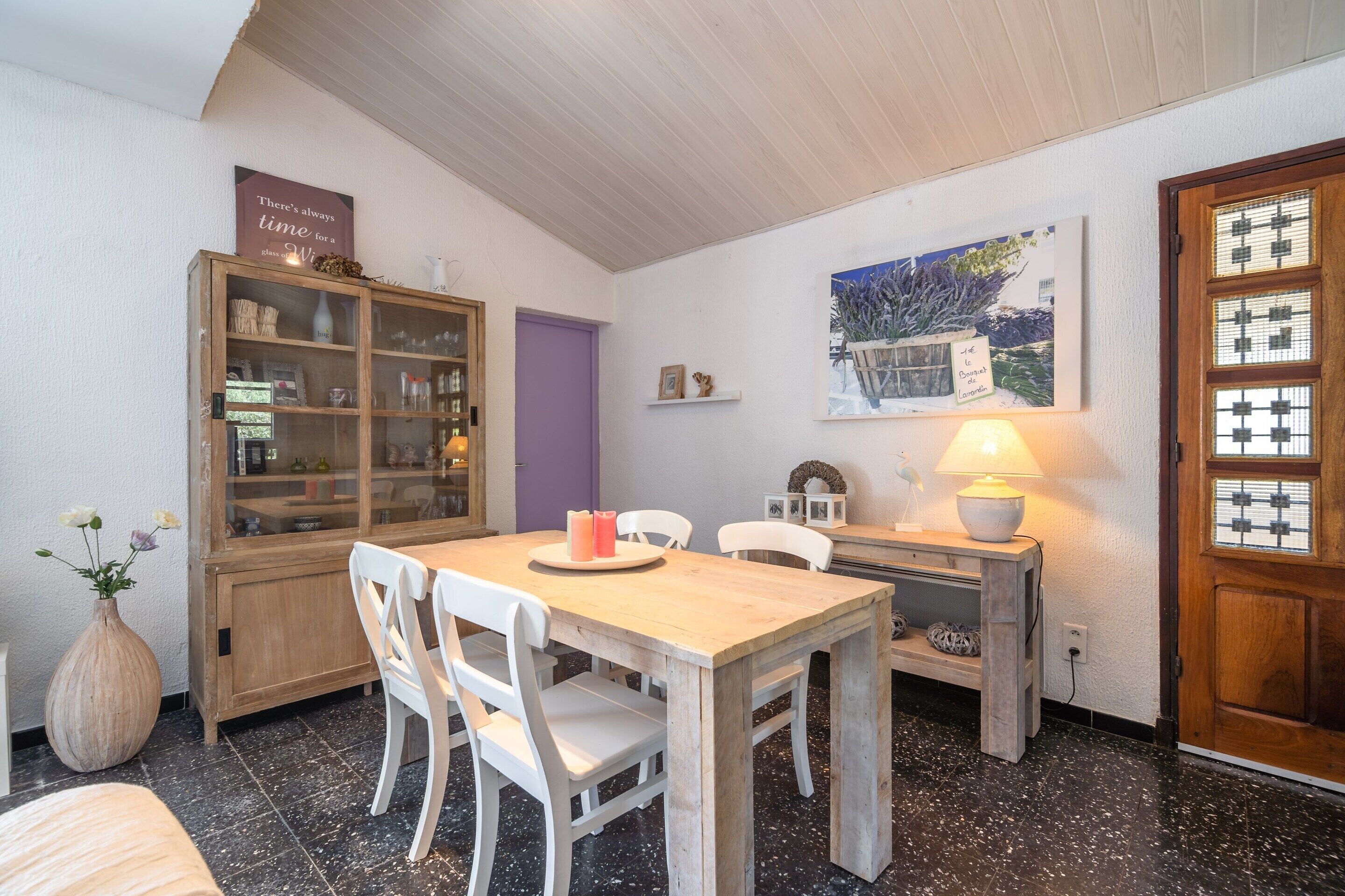 A dining area with a wooden table and chairs, a cabinet with glass doors, a purple door, and a wall-mounted painting.
