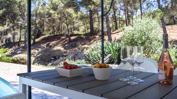 A table with food, glasses, and a bottle in a covered outdoor area with a pool and trees in the background. - Villa in Pouzols With Private Pool (Pouzols-Minervois)