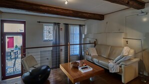 Ein Wohnzimmer mit Couch, Sessel und einem Holztisch. Es gibt ein Fenster mit Vorhängen, eine Tür, die nach draußen führt, und eine Heizung.