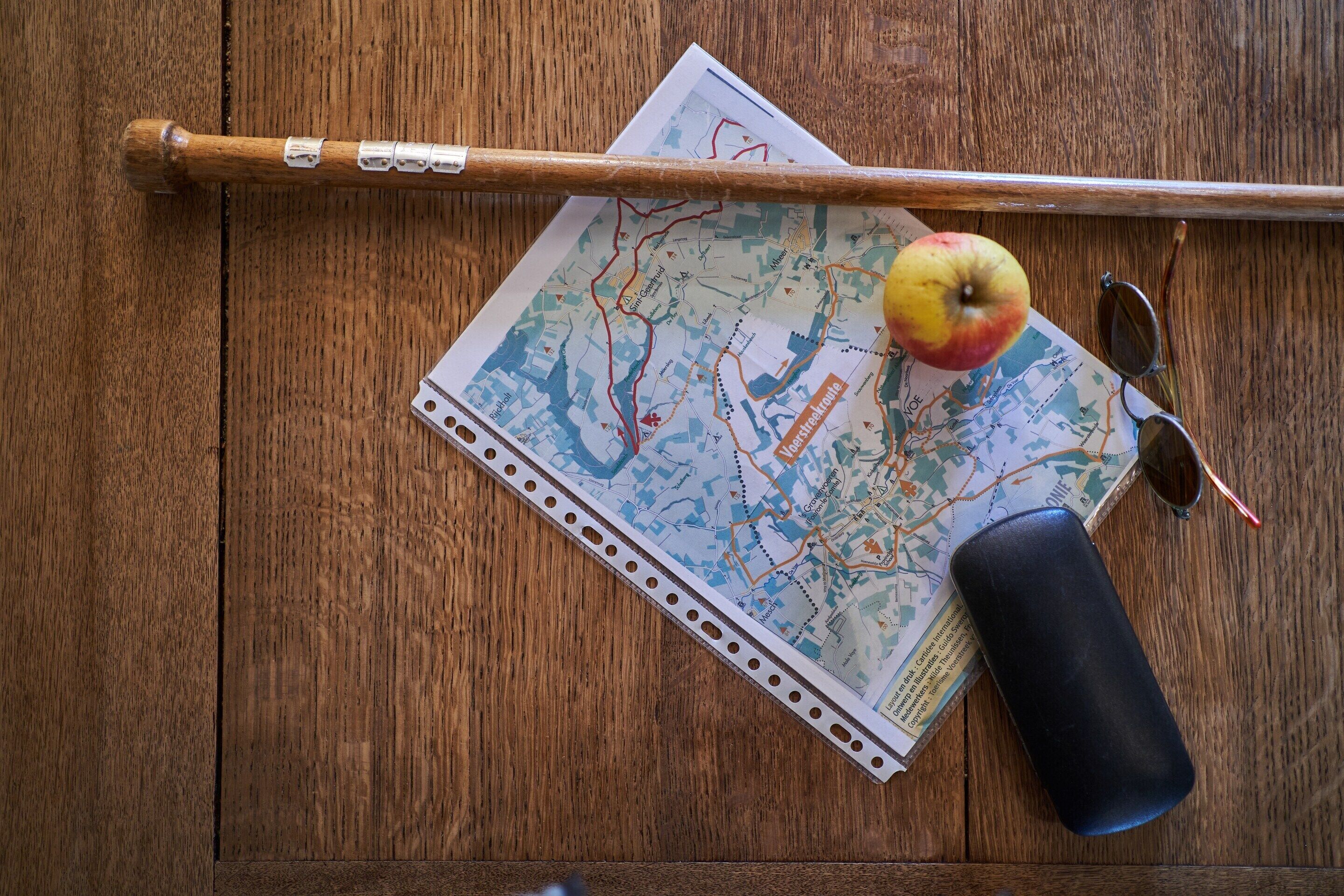 A wooden table with a map, a walking stick, a pair of sunglasses, and an apple.
