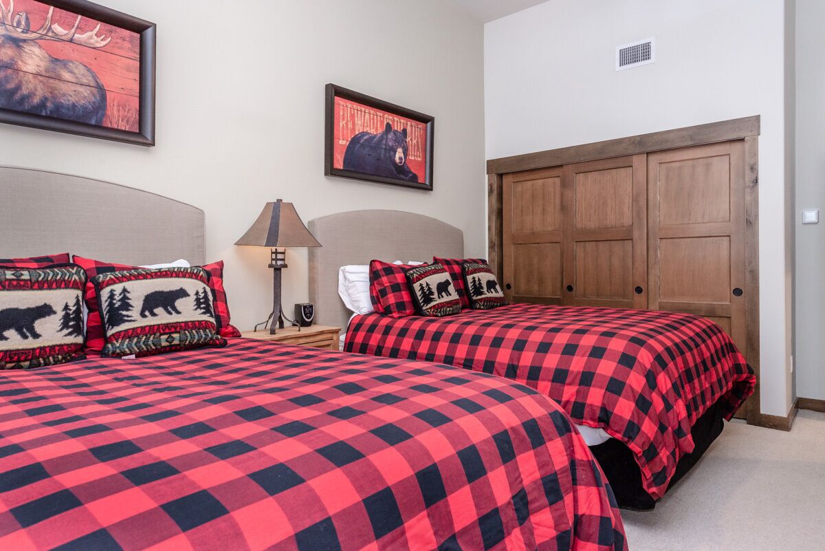 A bedroom with two beds, each with red and black checkered bedding and bear-themed pillows. There are two framed pictures on the wall, a lamp on the nightstand, and a wooden door.