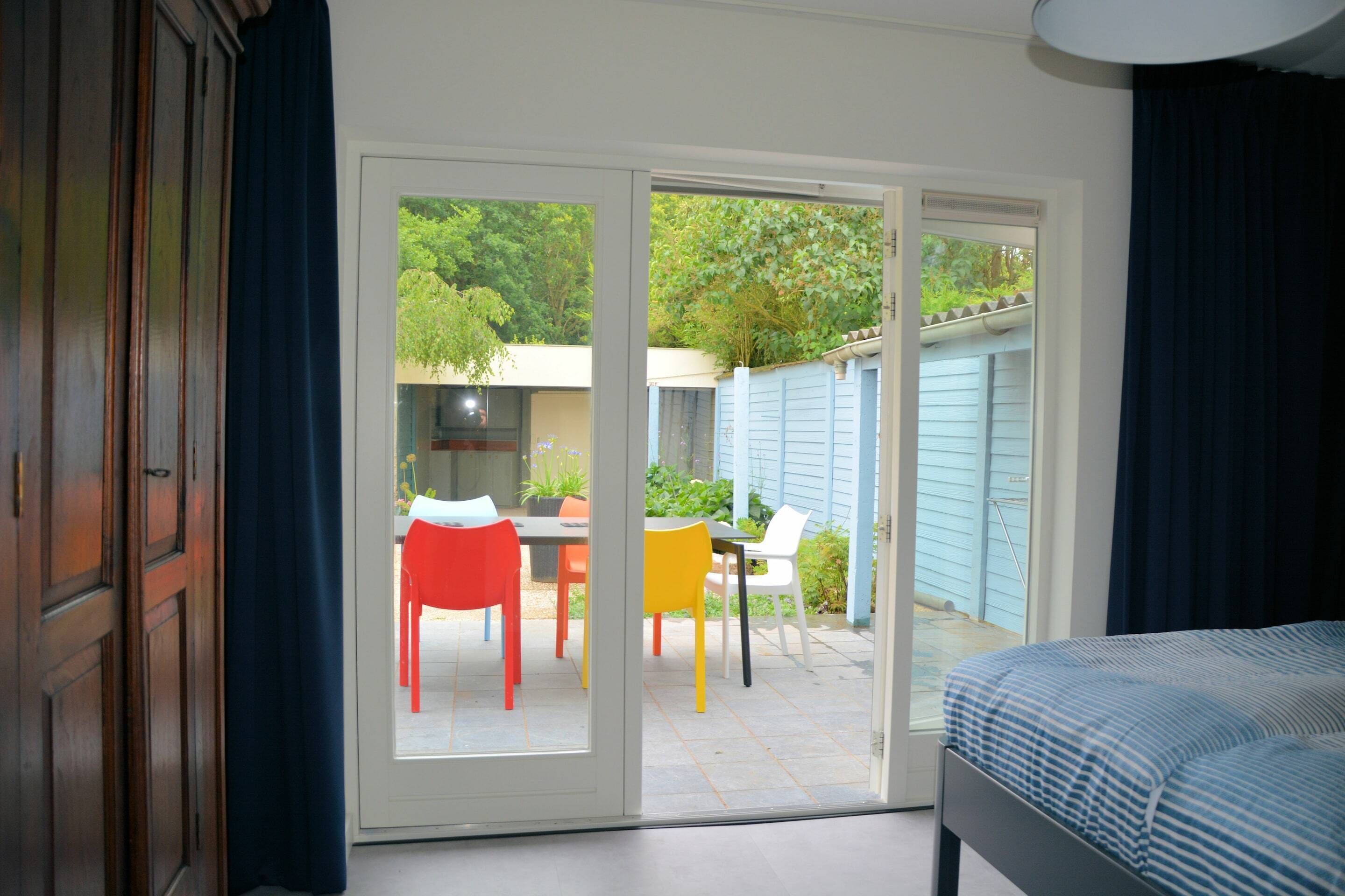 A bedroom with a bed, a sliding glass door leading to a patio with a table and chairs, and a view of greenery outside.