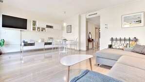 Un salon moderne avec un canapé, une table basse et un téléviseur fixé au mur. On y trouve également un coin repas avec une table et des chaises, ainsi qu’une cuisine avec un comptoir et des placards.