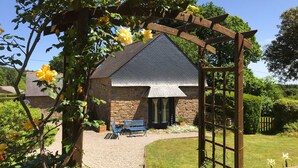 Ein Steinhaus mit geneigtem Dach, eine Holzpergola mit gelben Blüten und ein Kiesweg, der zu einer Bank führt.