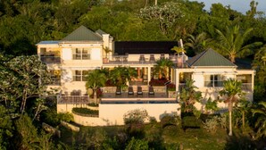 Una casa grande de dos pisos con piscina, rodeada de exuberante vegetación y palmeras.
