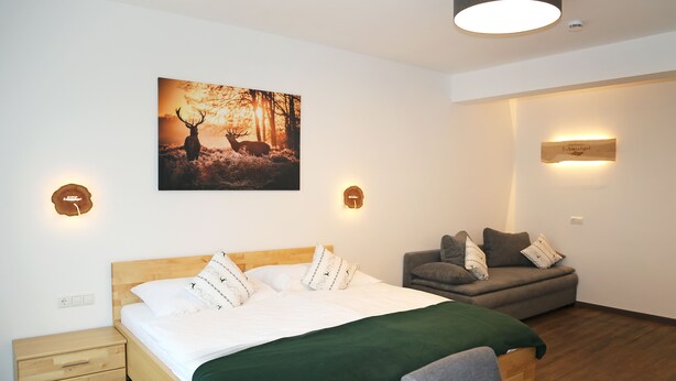 A bedroom with a bed, a nightstand, a sofa, and a wall-mounted light fixture. - Apartments Schmiedgut (Haus)