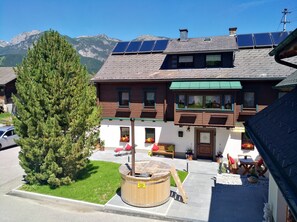A chalet with a wooden exterior, solar panels on the roof, a wooden hot tub, and a view of mountains.