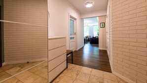 A hallway with white brick walls, a wooden floor, and a door leading to a living area with a dark-colored sofa and a hanging light fixture. - Victus Apartamenty - Walor 2 (Sopot)
