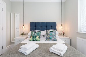 A neatly made bed with a blue headboard, two bedside tables with lamps, and a window with blinds.