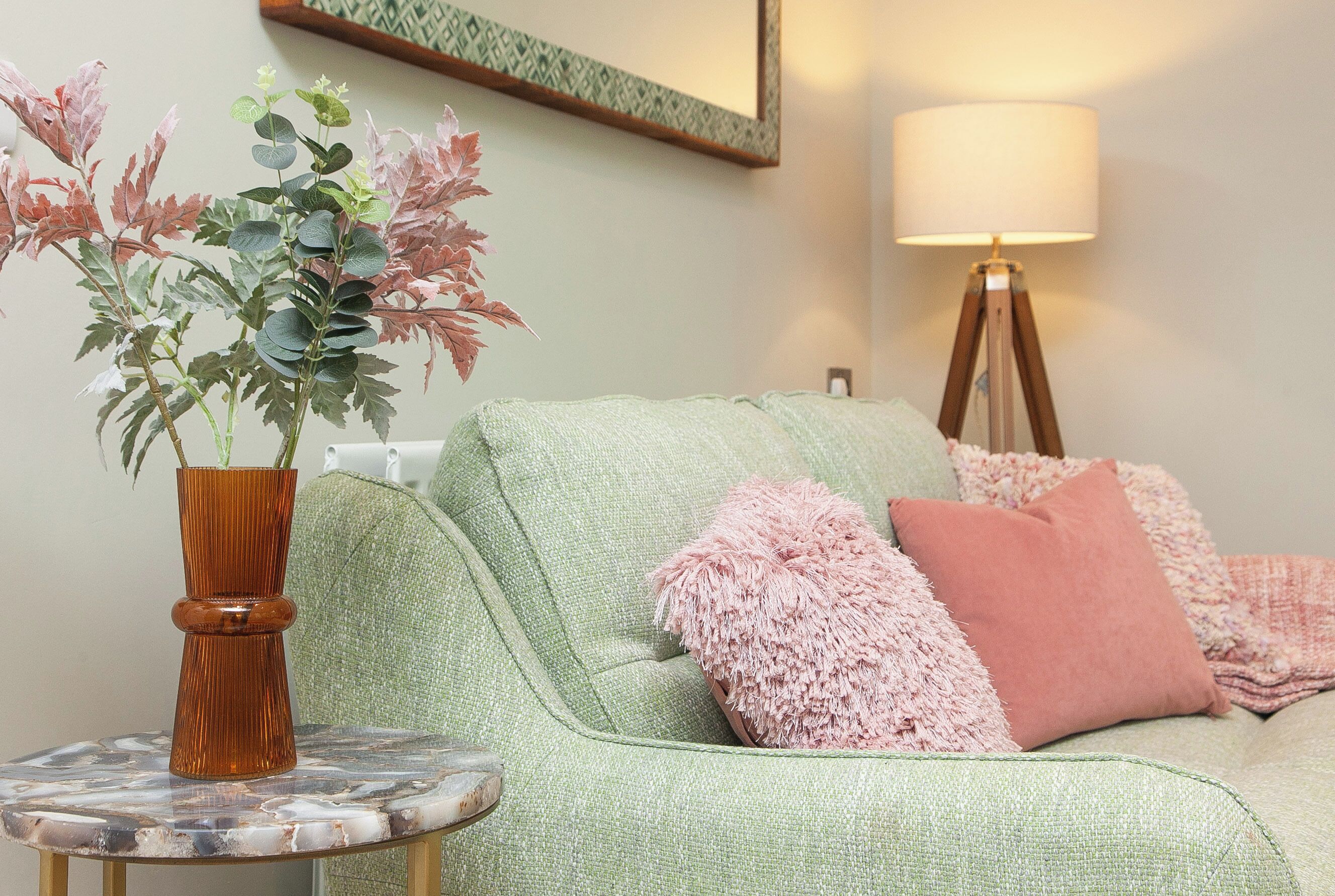 A green sofa with pink and white throw pillows, a small round table with a vase of flowers, and a floor lamp.