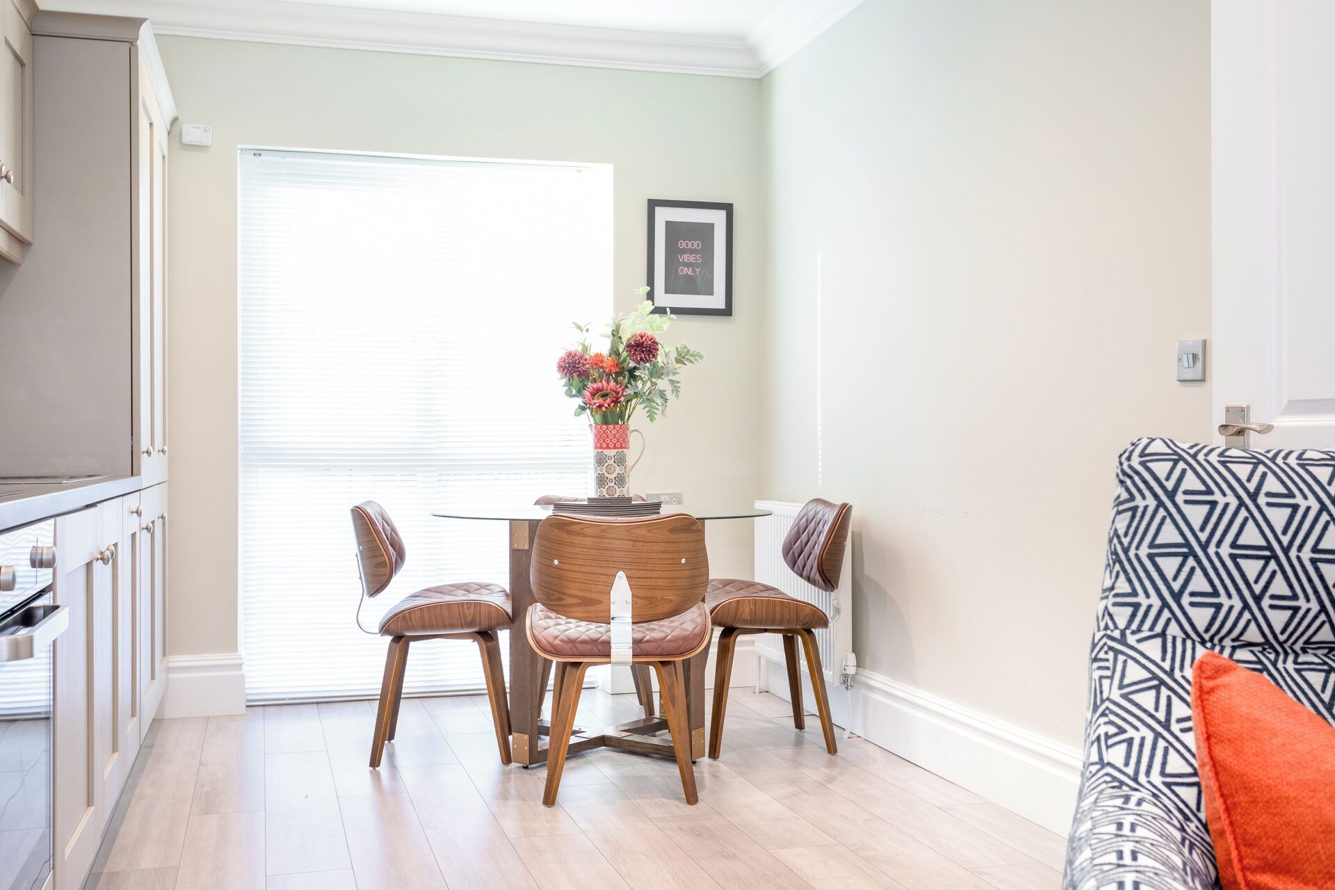Un comedor con una mesa y cuatro sillas, un florero con flores, un cuadro en la pared y un sofá estampado.
