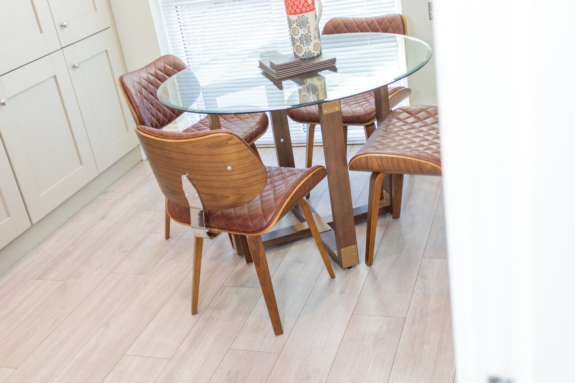 A dining area with a glass-top table, four wooden chairs with cushioned seats, and a patterned vase on the table.