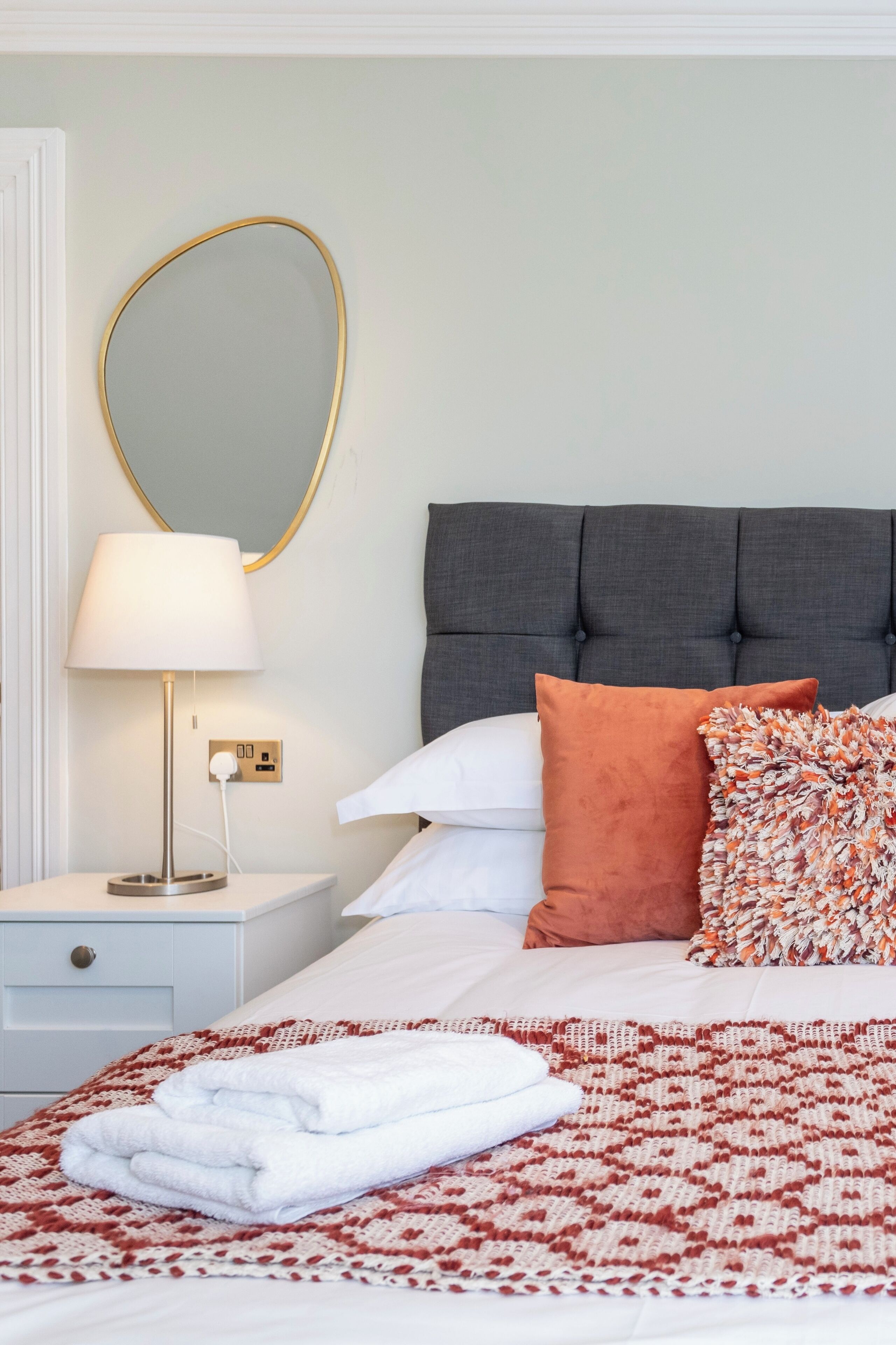 A neatly made bed with a patterned blanket, a bedside lamp, and a mirror on the wall.