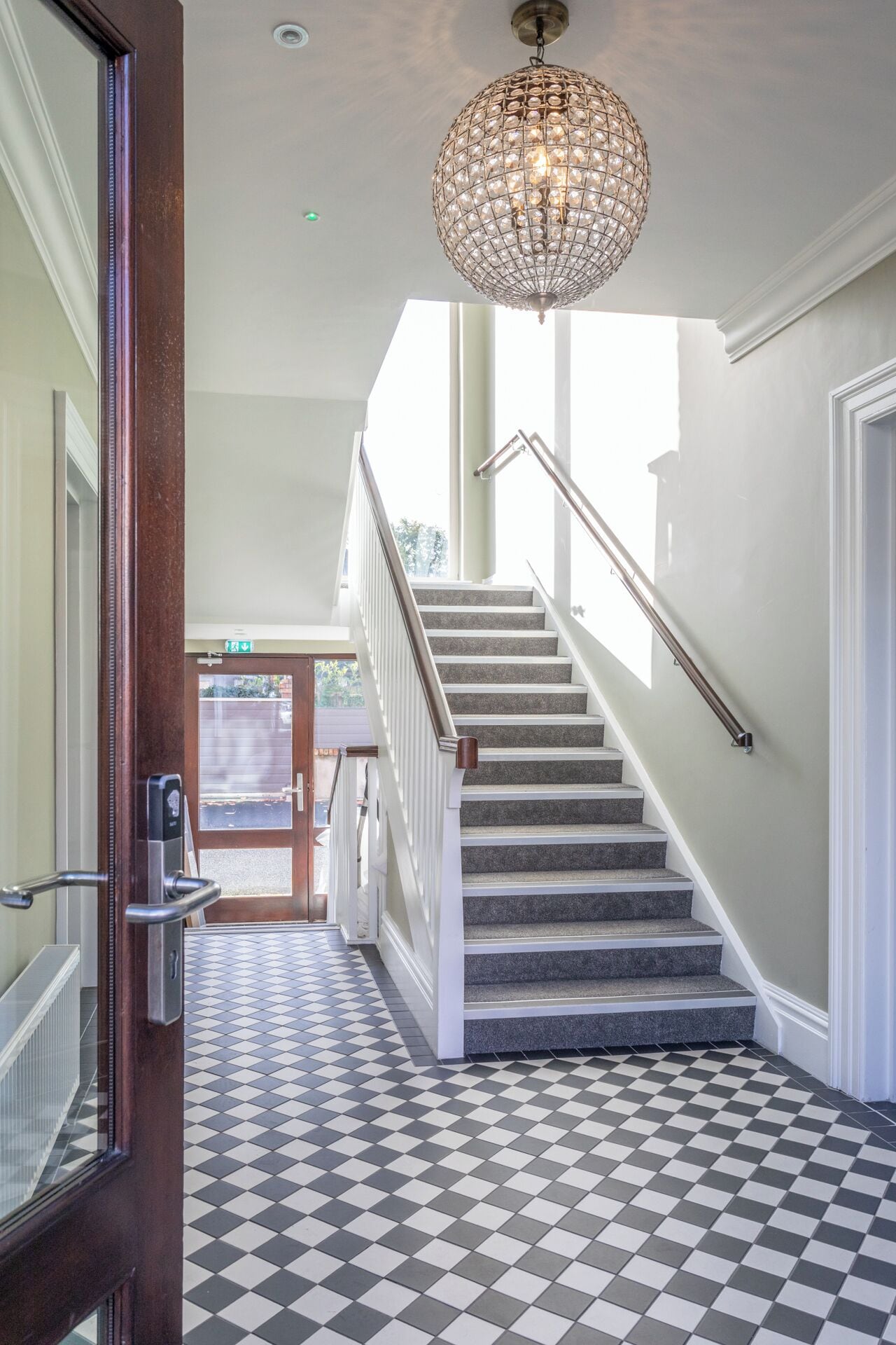 A staircase with a checkered floor leading to a landing, a chandelier, and a door with a security device.