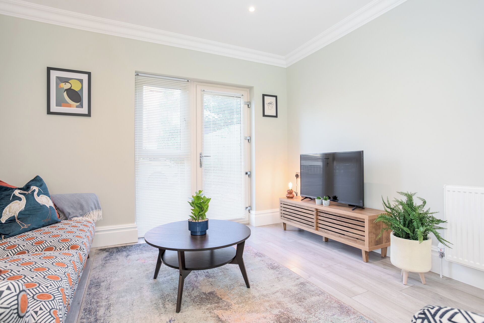 Ein Wohnzimmer mit einer Couch, einem runden Tisch mit einer Pflanze, einem Fernseher auf einem Holztisch und einer Heizung.