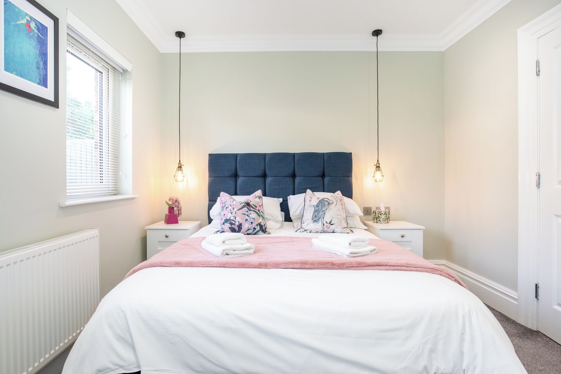 A bedroom with a large bed, two bedside tables, a headboard, and a window with blinds.