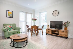 A living room with a green sofa, a wooden coffee table, a dining area with wooden chairs, a TV on a wooden stand, and a clock on the wall.