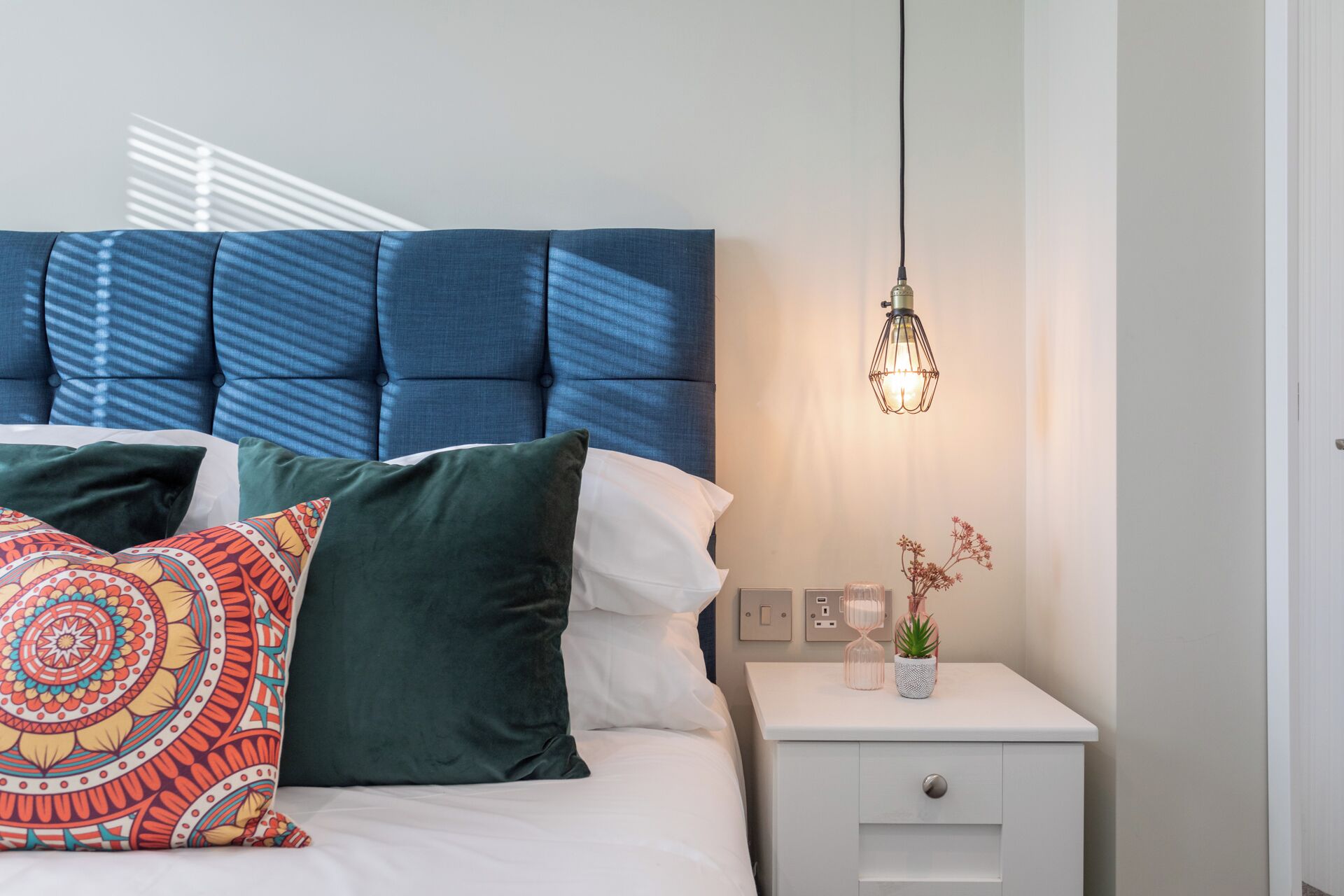 A neatly made bed with a blue upholstered headboard, white and green pillows, and a decorative pillow with a colorful pattern. A bedside table with a small potted plant and a wall-mounted light fixture is visible.