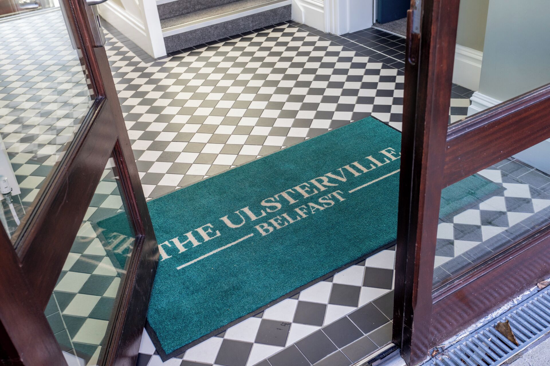 A green doormat with "THE ULSTERVILLE BELFAST" printed on it, placed at the entrance of a room with a checkered floor pattern.