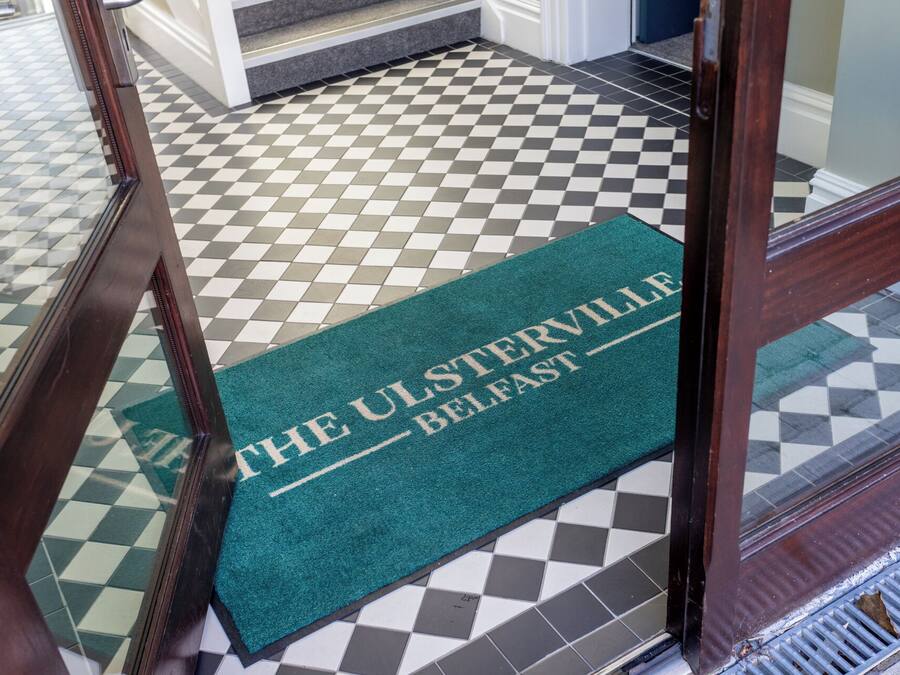 A green doormat with "THE ULSTERVILLE BELFAST" printed on it, placed at the entrance of a room with a checkered floor pattern.