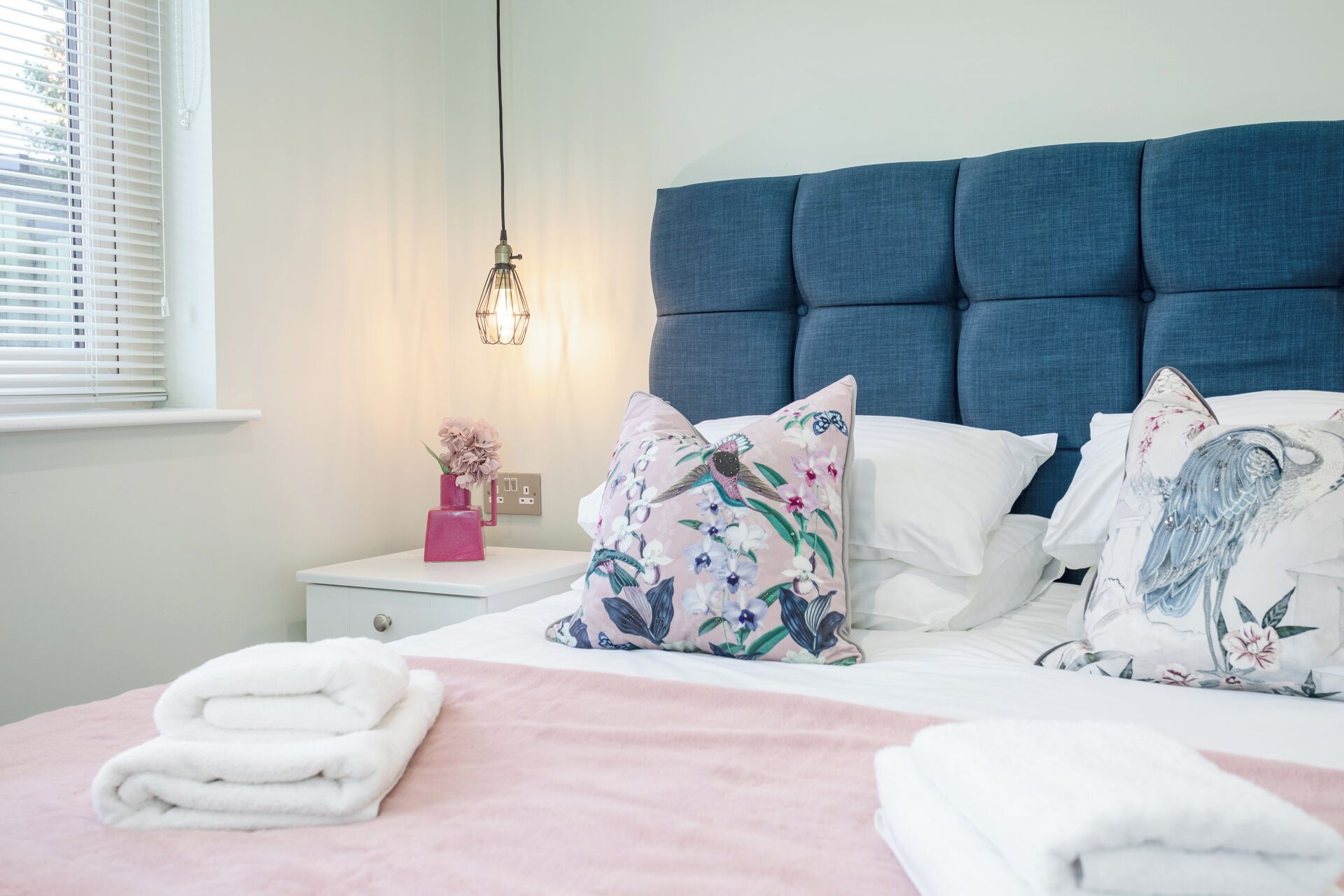 Ein ordentlich gemachtes Bett mit floralen Kissen, ein Nachttisch mit einer rosa Vase und eine hängende Leuchte.