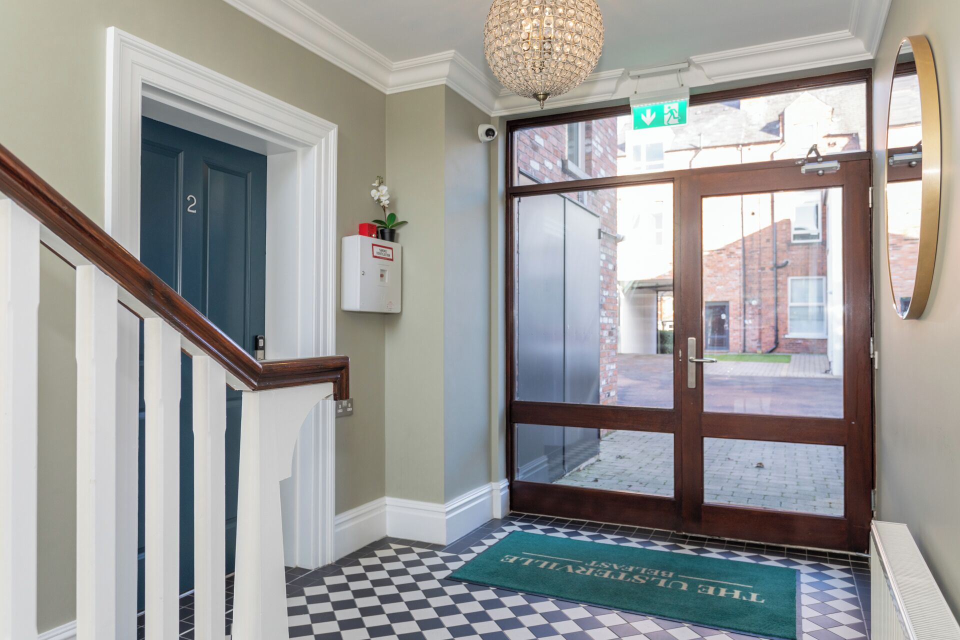 L’entrée d’un hôtel, avec un paillasson à carreaux, une porte surmontée d’un panneau vert indiquant la sortie de secours, et un interphone de sécurité fixé au mur.