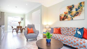 Een woonkamer met een gestreept bankje, een ronde salontafel en een fauteuil. Er hangt een schilderij aan de muur en er is een eethoek op de achtergrond.