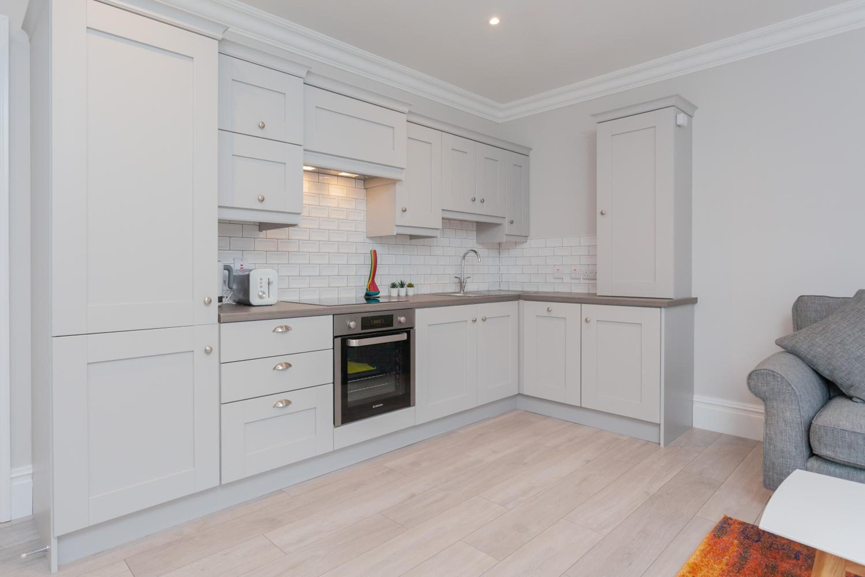A modern kitchen with white cabinetry, a built-in oven, and a grey sofa in the adjacent living area.