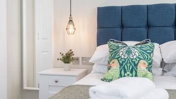 Un lit bien fait, avec une tête de lit bleue, une lampe de chevet, une petite table de chevet ornée d’une plante et un oreiller à motifs verts et oranges.