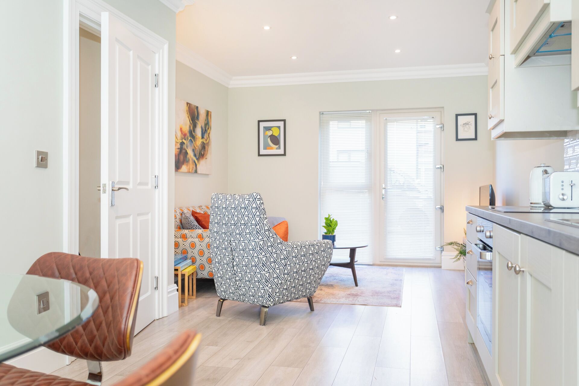 A modern living room with a patterned armchair, a glass table, and a view of the kitchen.