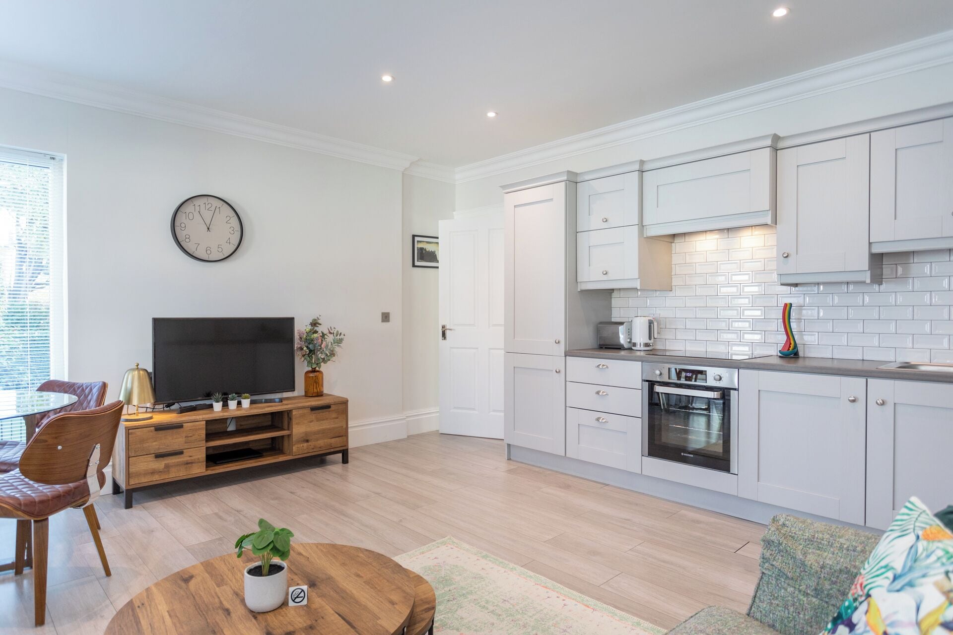 Una cucina moderna con mobili bianchi, un forno integrato e un microonde. C'è un'area pranzo con un tavolo e sedie in legno, e un televisore su un mobile in legno.