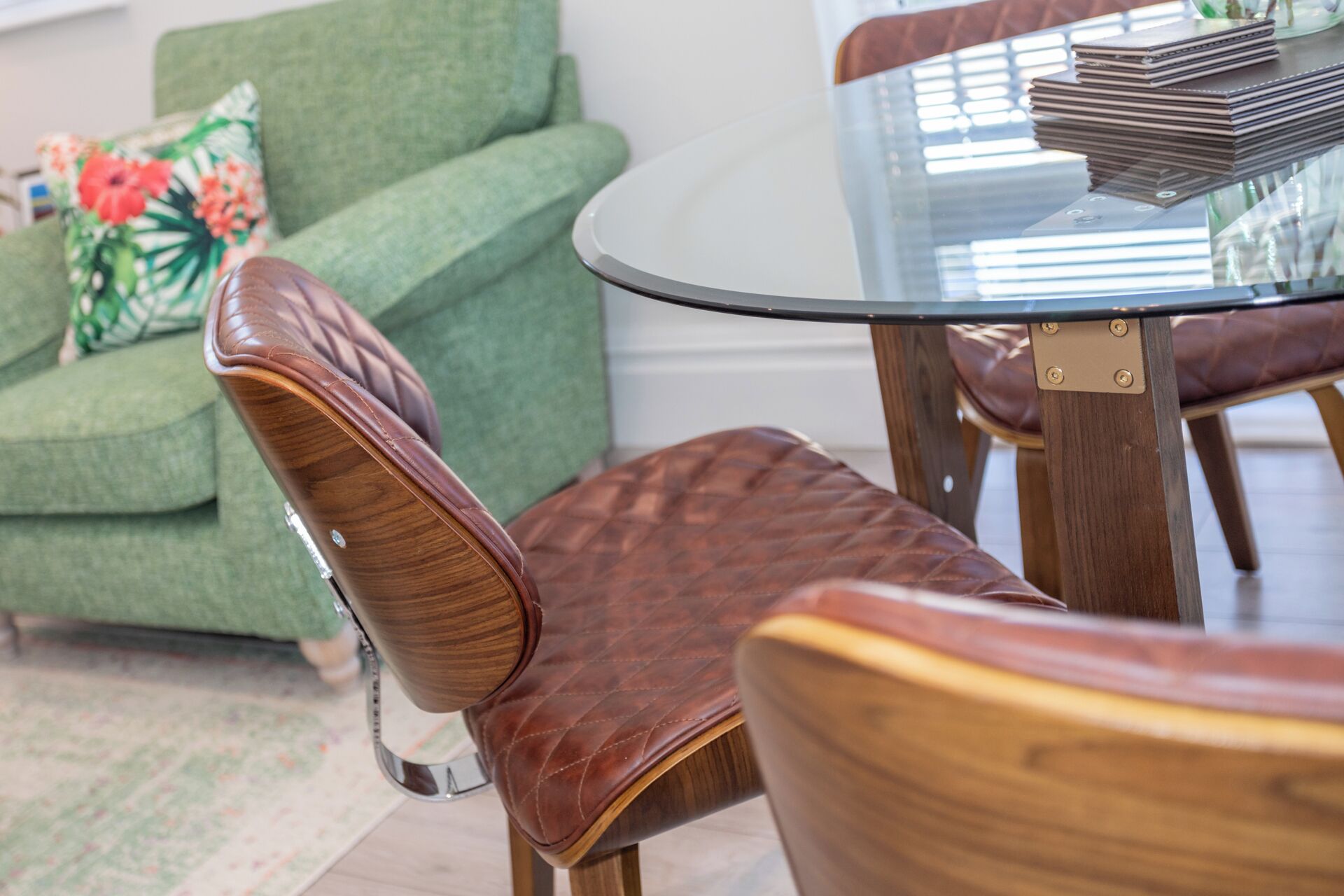 A dining area with a glass table and leather chairs, a green sofa with floral cushions, and a stack of books on the table.