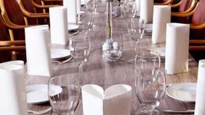 Uma longa mesa de jantar posta para um evento formal, com guardanapos brancos, copos de vidro transparente e um vaso com rosas cor-de-rosa.