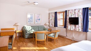 Quarto de hotel com um sofá verde, cadeiras de madeira, uma mesa de madeira, uma cama, uma televisão e duas imagens emolduradas na parede.