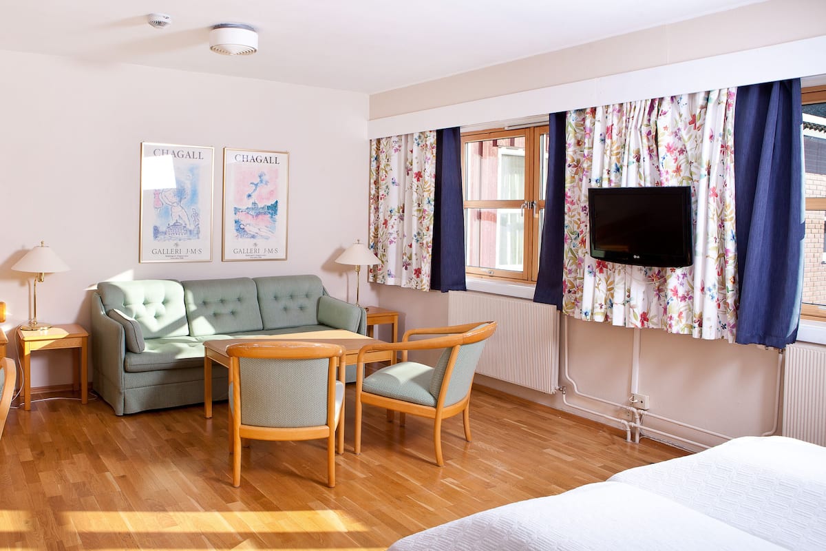 A hotel room with a green sofa, wooden chairs, a wooden table, a bed, a TV, and two framed pictures on the wall.