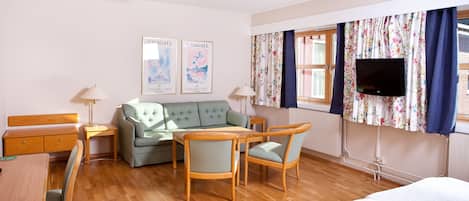 A hotel room with a green sofa, wooden chairs, a wooden table, a bed, a TV, and two framed pictures on the wall.