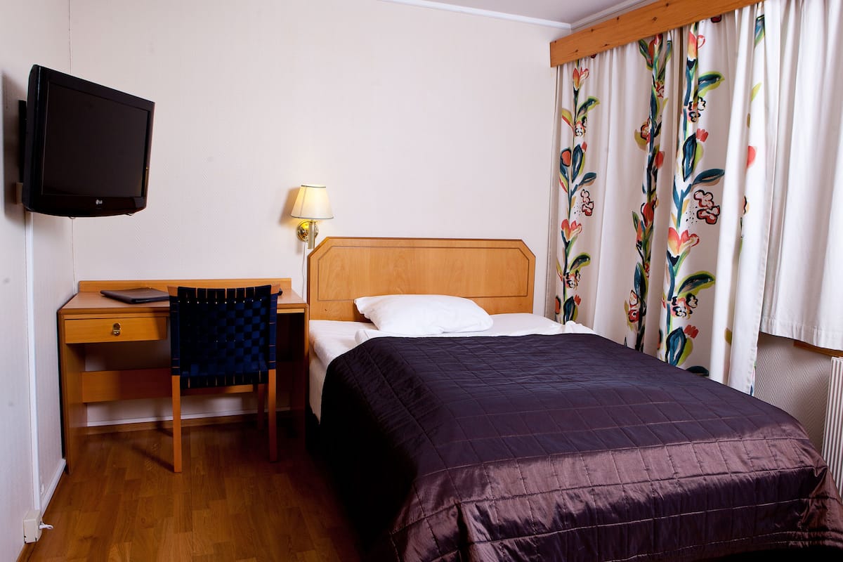 A hotel room with a wooden bed, a desk with a chair, a television, and a window with floral curtains.