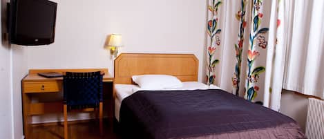 Habitación de hotel con una cama de madera, un escritorio con una silla, un televisor y una ventana con cortinas florales.