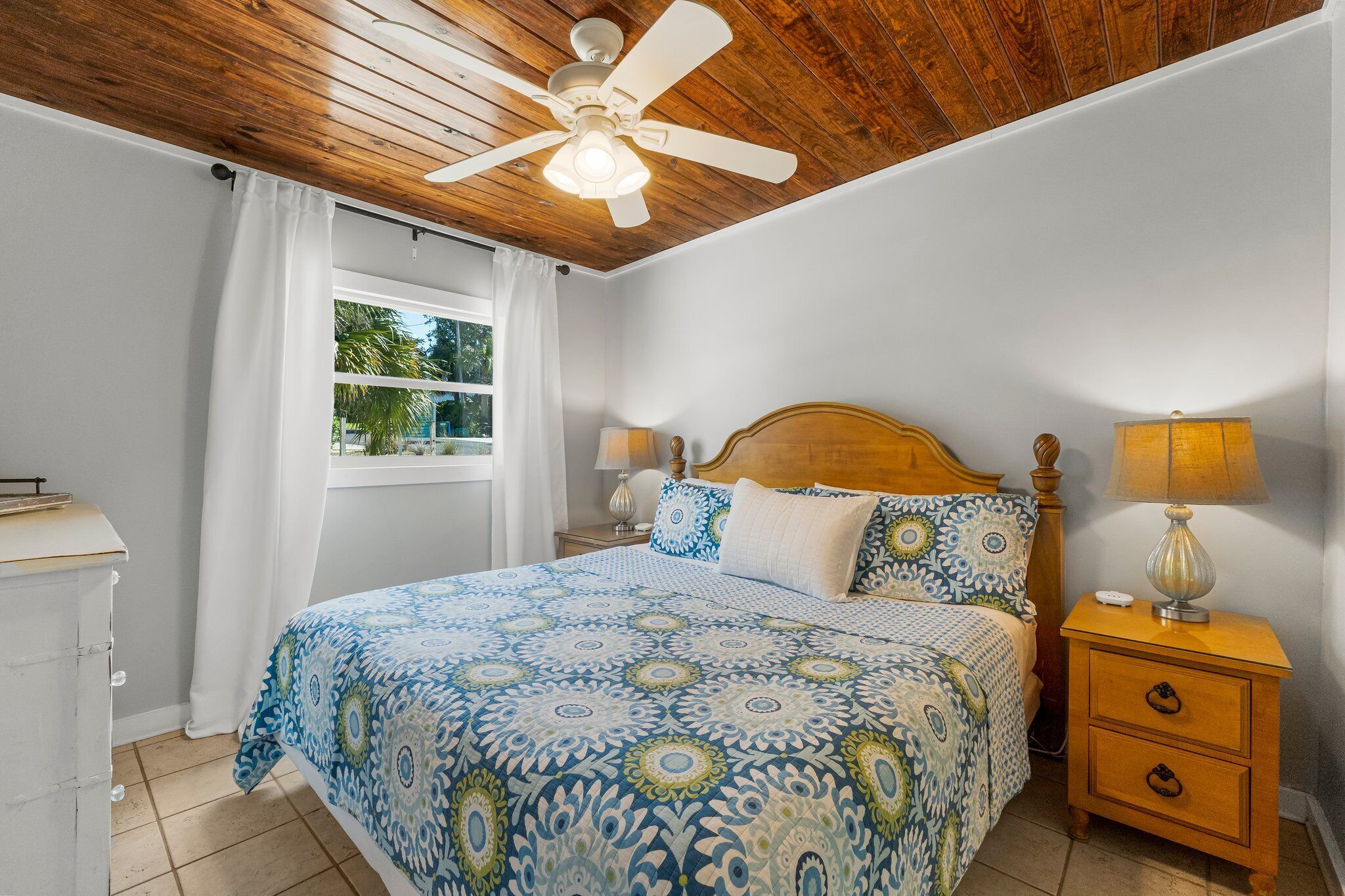 A bedroom with a wooden ceiling, a bed with patterned bedding, two bedside lamps, and a window with curtains.