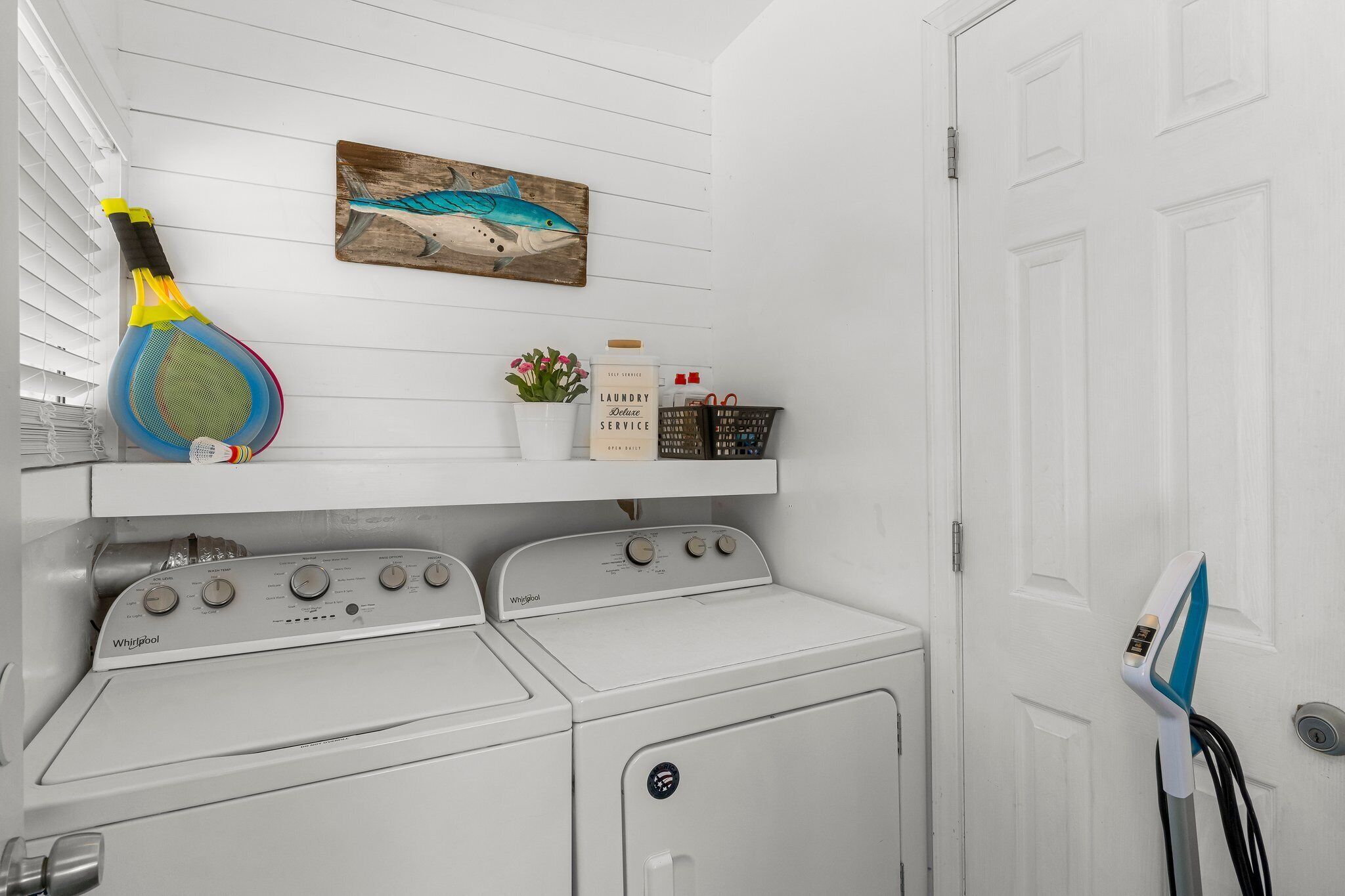 A laundry area with a Whirlpool washer and dryer, a shelf with a painting, and a basket of laundry supplies.